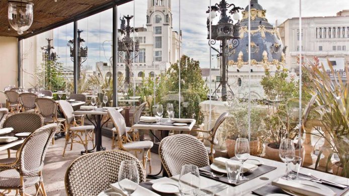 Glass-enclosed terrace with rattan furniture and potted plants, overlooking rooftops