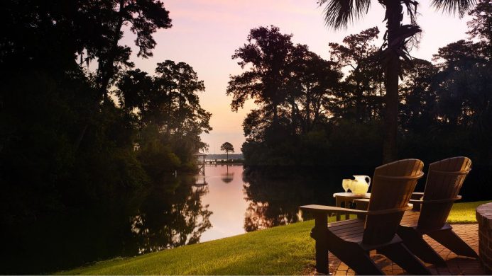 Adirondack chairs on a green lawn on the riverfront under a pink sky at Montage Palmetto Bluff