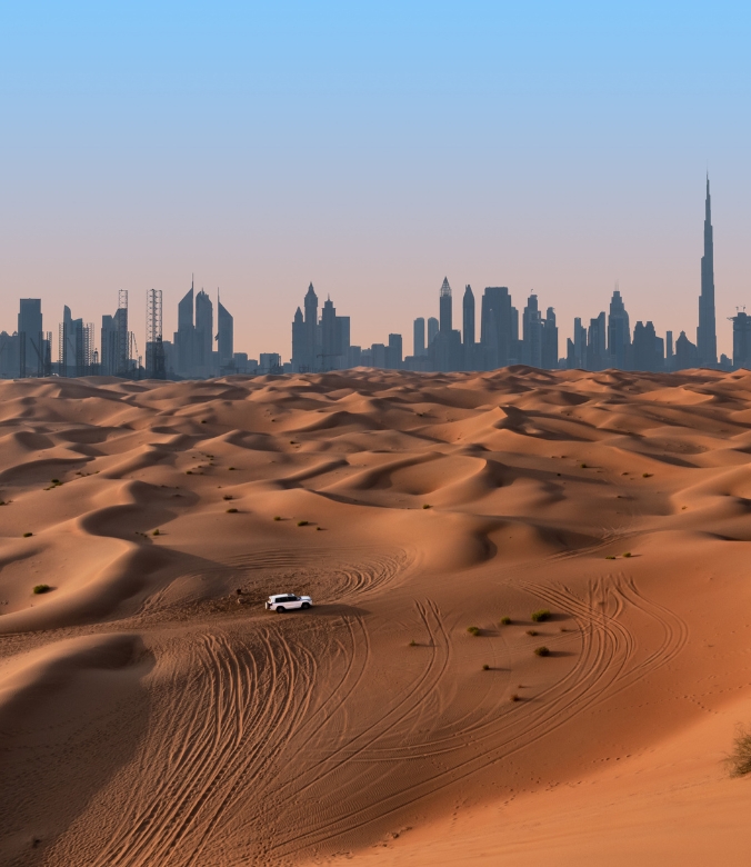 Day trip - a car driving through desert dunes in front of the Dubai skyline
