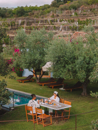 Where to stay in Sicily travel diary - two people eating breakfast surrounded by grass and trees next to a pool at Monaci delle Terre Nere Etna Wine Resort